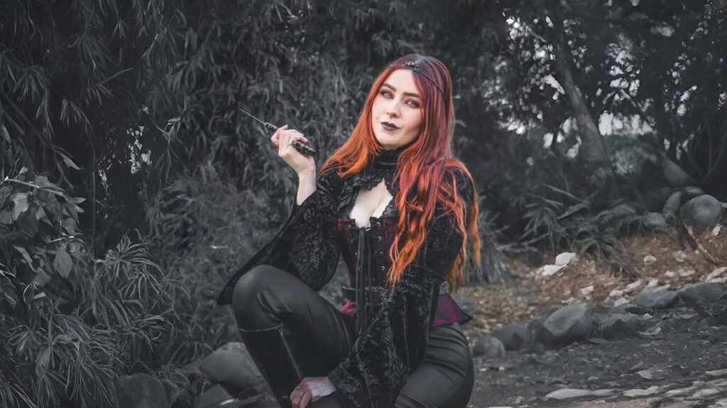 A woman with long red hair poses confidently in a forest setting, wearing a black velvet corset top with lace detailing and flared sleeves, paired with sleek leather pants. The outfit, inspired by Renaissance fashion, exudes a dark, mystical aesthetic reminiscent of gothic or fantasy styles.