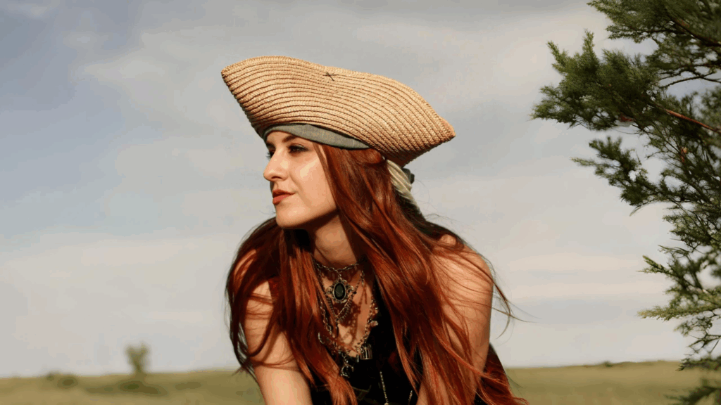 Red-haired woman in piratecore outfit wearing a tricorn hat and layered necklaces, posing outdoors with a vintage aesthetic.