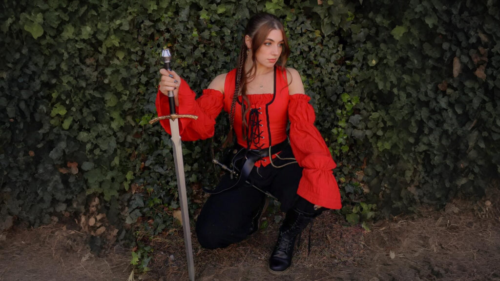 A woman dressed in a vibrant red pirate-style off-shoulder blouse and black trousers, kneeling in front of a leafy wall while holding a medieval-style sword.