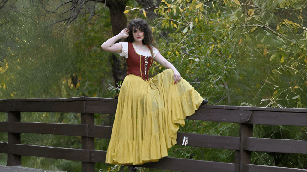 Woman wearing a Renaissance-style outfit with a yellow skirt and corset top, standing on a wooden bridge surrounded by green forest.