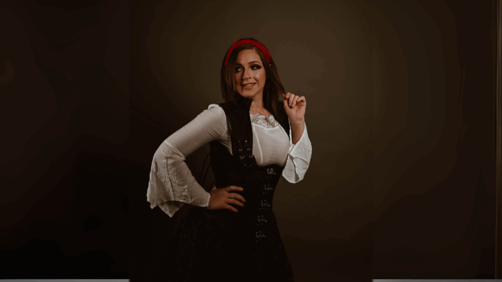 A woman wearing a black steampunk corset over a white pirate shirt with bell sleeves, styled with a red headband, posing confidently against a dark background. Fantasy piratecore fashion by The Pirate Dressing.