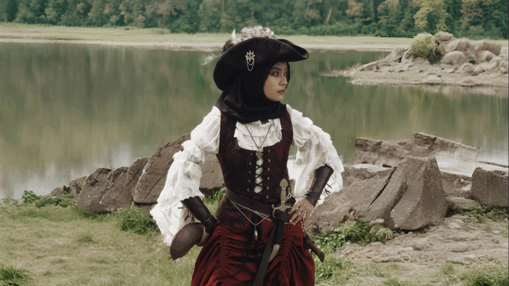 Woman wearing a black pirate hat, white ruffled pirate shirt, and burgundy corset with sword, standing by a lake in fantasy-themed pirate clothing