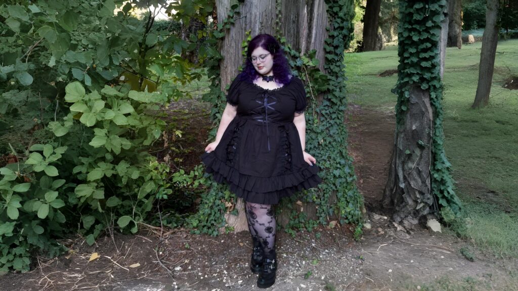 Woman in a black Lolita Goth dress with lace details, posing outdoors in a forest setting.