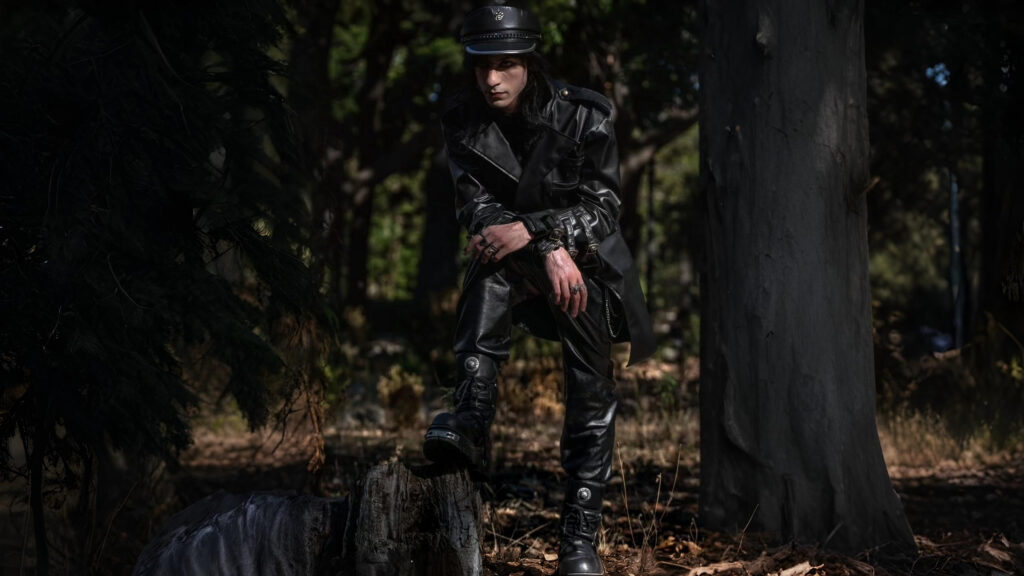 Man in black steampunk trench coat and leather boots posing outdoors in a dark, forest setting.