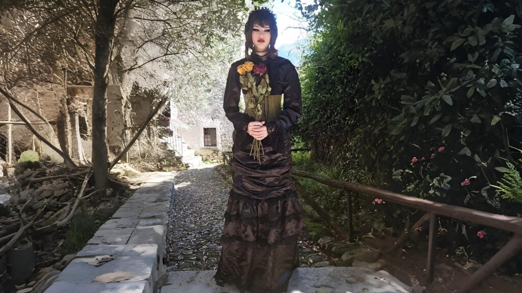 Model in The Wickfield 3-piece steampunk ensemble, wearing a black Victorian-style skirt, blouse, and corset while holding roses in a rustic garden setting.