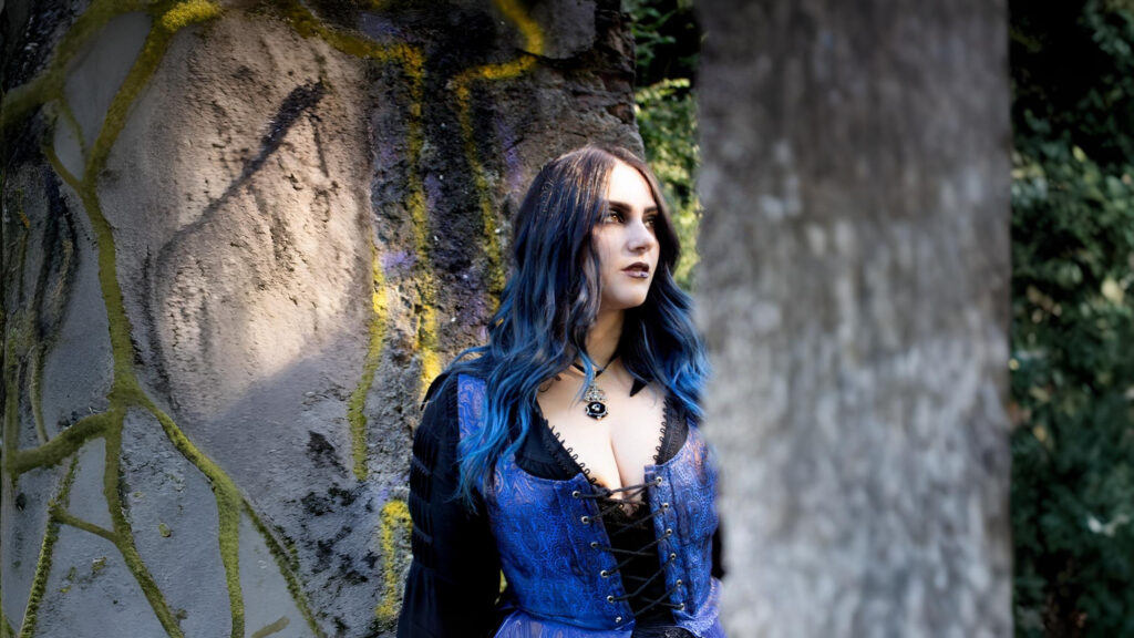 Woman in a blue corset-style Gothic outfit standing near mossy stone pillars, embodying dark romantic Gothic fashion.