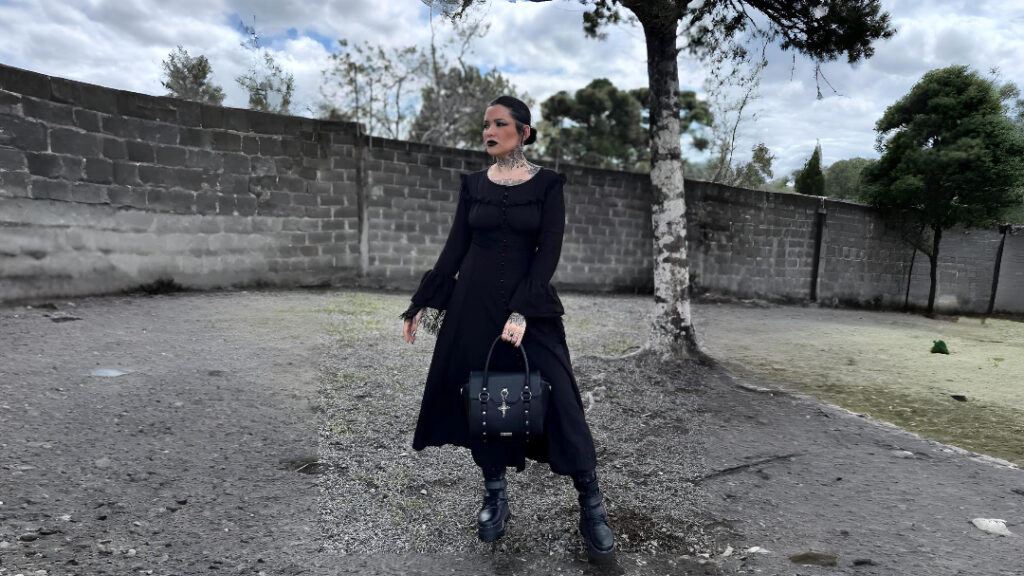 Gothic woman in a long black dress with bell sleeves, black boots, and a structured gothic handbag styled outdoors.