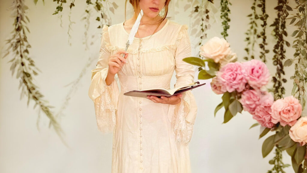 Woman in a cream medieval dress holding a quill and book surrounded by hanging vines and pink roses.