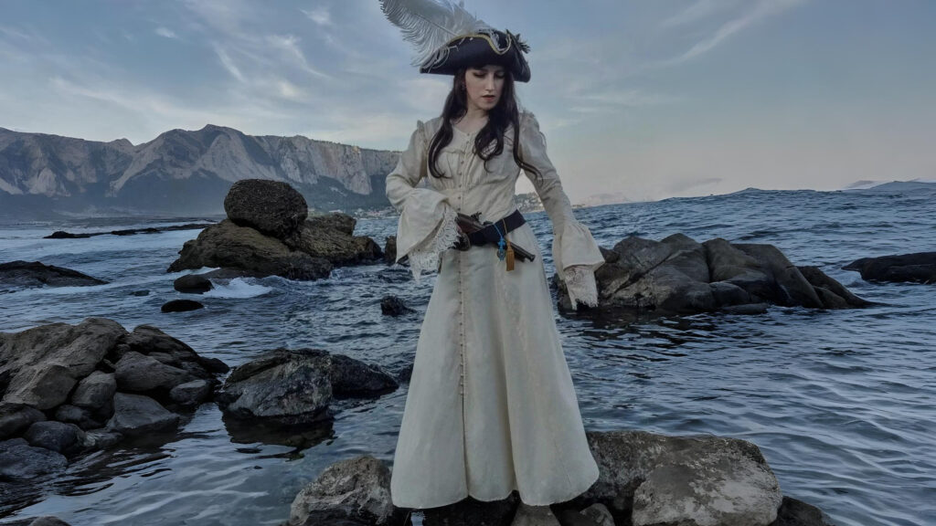A woman dressed in an elegant cream Renaissance pirate coat with lace sleeves and a feathered tricorn hat stands on seaside rocks, embodying a regal yet minimalist pirate aesthetic from The Pirate Dressing.