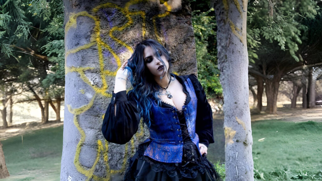 Model wearing a blue steampunk corset with black lace-up detailing from The Pirate Dressing, styled with a black Victorian blouse and gothic accessories, posing against a rustic stone wall.