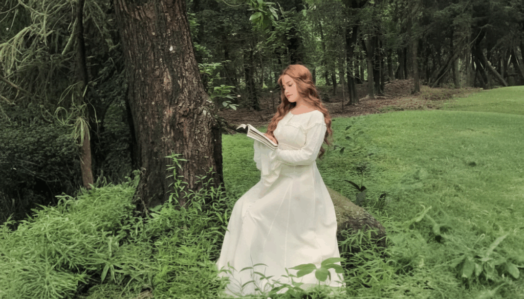 A woman wearing a soft white medieval-style dress sits beside a large tree in a lush green forest, reading a book with calm, serene expression.