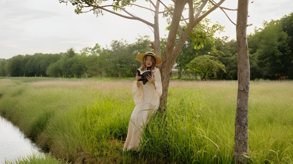 Woman in a long cream renaissance-style dress reading a book under a tree in a green meadow.