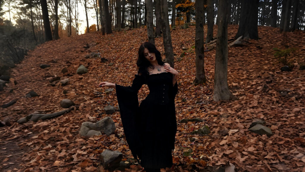 Woman wearing a black medieval-style dress with flowing sleeves standing in an autumn forest covered in fallen leaves