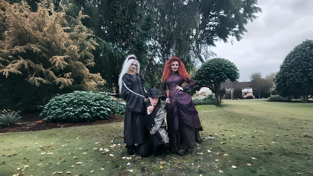 Three models dressed in steampunk Victorian gothic outfits posing outdoors in a lush garden setting, showcasing lace-detailed gowns and a patterned steampunk coat by The Pirate Dressing.