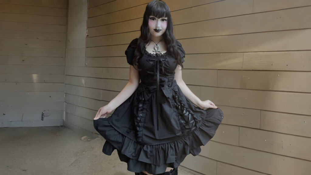Person wearing a black gothic lolita dress with layered ruffles, lace details, and soft curls, standing against a wooden background.