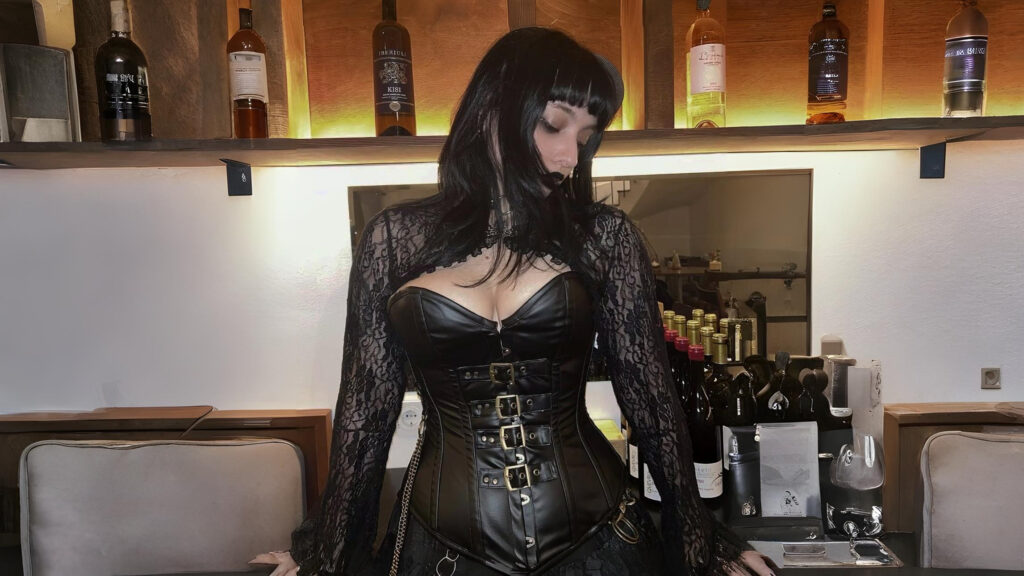 Woman wearing a black leather steampunk corset with lace sleeves, styled in gothic steampunk fashion at a bar setting.