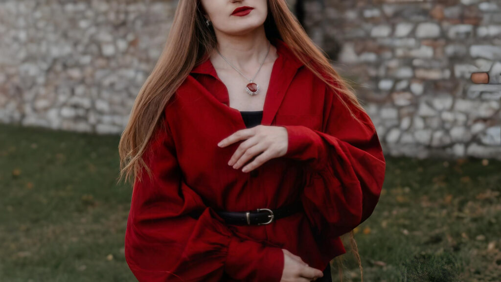 A woman wearing a deep red pirate-style shirt with voluminous sleeves, belted at the waist, standing outdoors near a stone wall. She has long straight hair, red lipstick, and a silver pendant necklace.