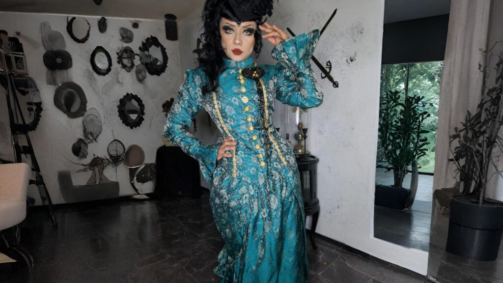 A model wearing a teal Victorian steampunk dress with gold buttons and lace detailing, posing indoors with one hand on her hip and the other touching her hat, surrounded by vintage wall décor.