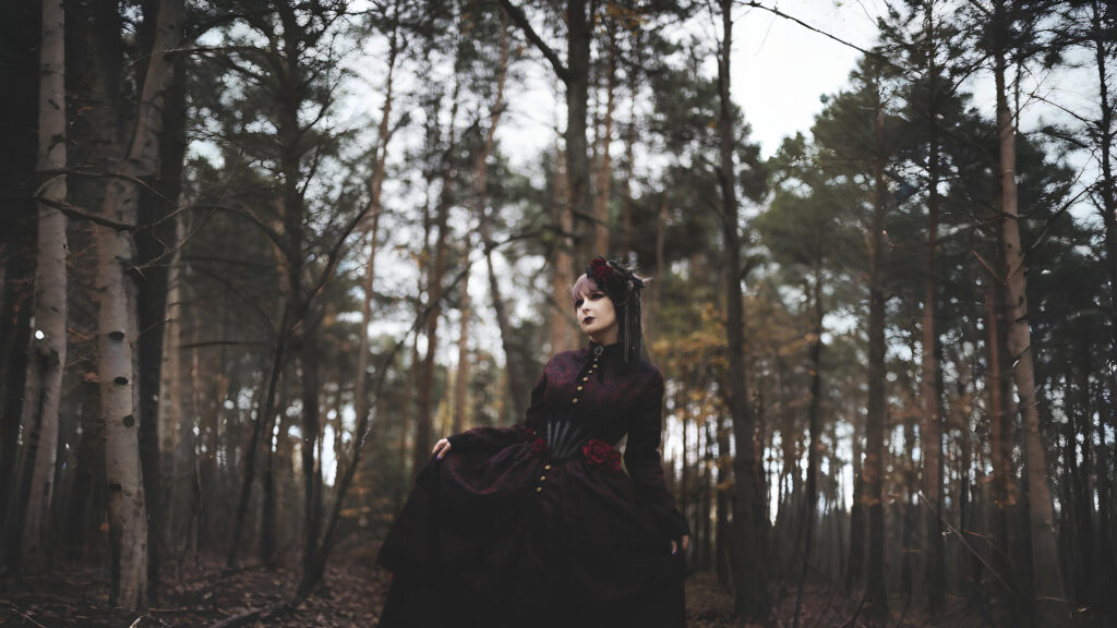 A woman in a dark burgundy gothic Renaissance-style dress standing in a forest, holding her skirt, with dramatic makeup and a floral headpiece