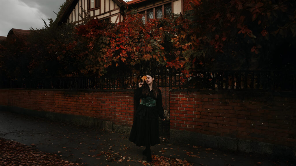 Woman wearing a dark green velvet corset over a black dress, standing beside a brick wall covered in autumn ivy leaves, inspired by historical and gothic fashion.