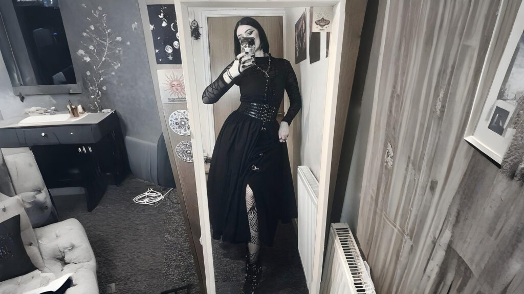 A person wearing a black gothic Renaissance-inspired outfit with a belted waist, mesh tights, and heeled boots, standing in front of a mirror in a softly lit room.