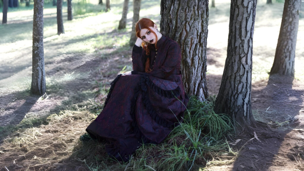 Woman wearing a dark Tudor-inspired historical dress sitting against a tree in a forest, evoking gothic and renaissance fashion aesthetics.